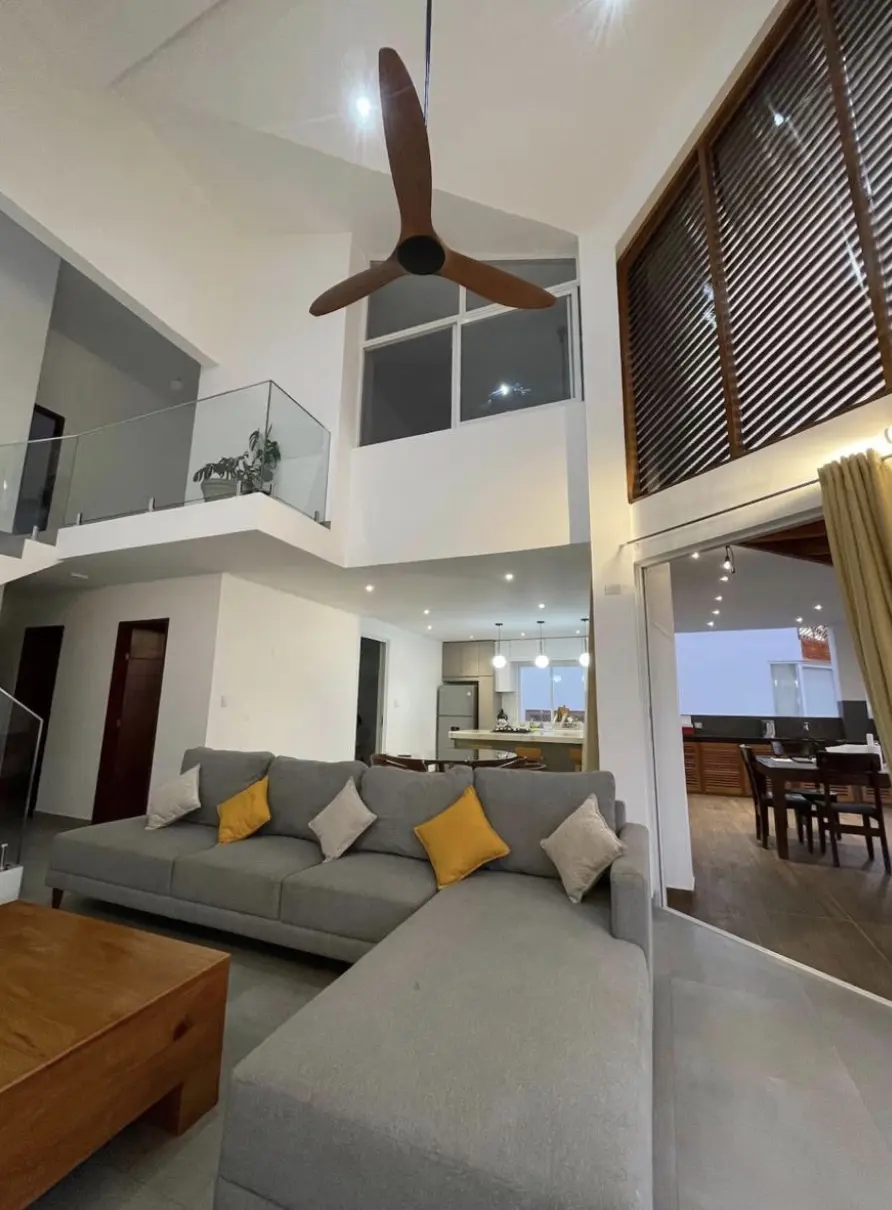 Sala de doble altura con sofá gris, ventilador de madera y vista a la cocina